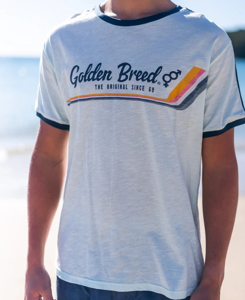 A person wearing a Retro Ringer Tee | Ice with navy blue trim on the collar and sleeves, featuring a Golden Breed logo and colorful diagonal stripes, standing by the beach on a sunny day.