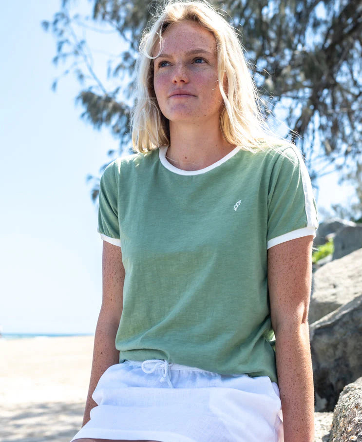 Woman wearing a Retro Ringer Tee | Moss with white trim, sitting outdoors near the beach on a sunny day with trees in the background.