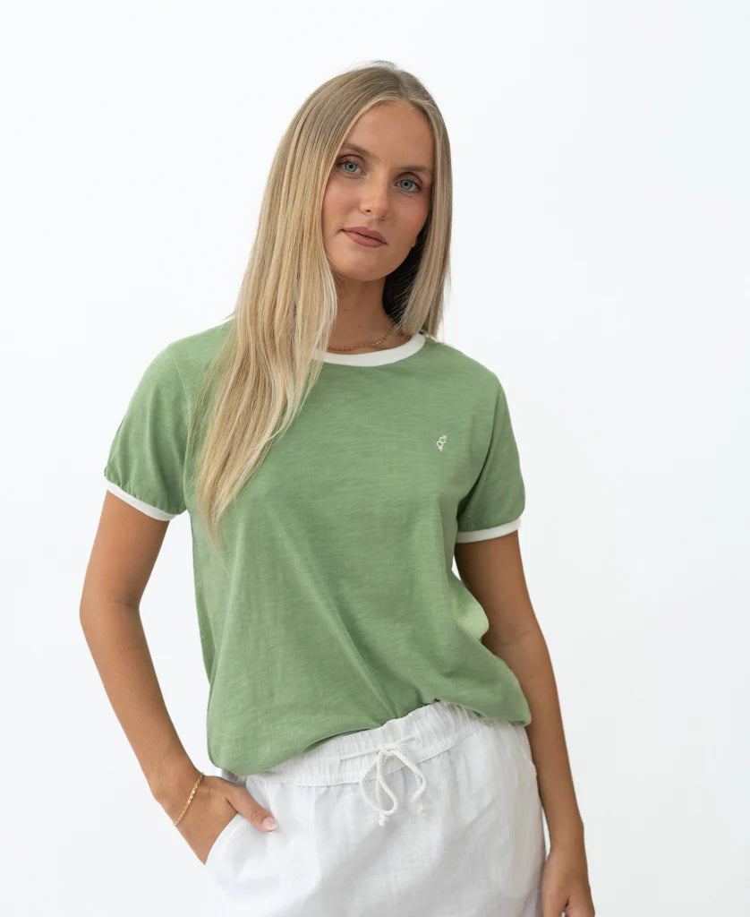 Woman wearing a green Retro Ringer Tee | Moss with white trim on sleeves and collar, paired with white drawstring pants, posing against a plain white background.