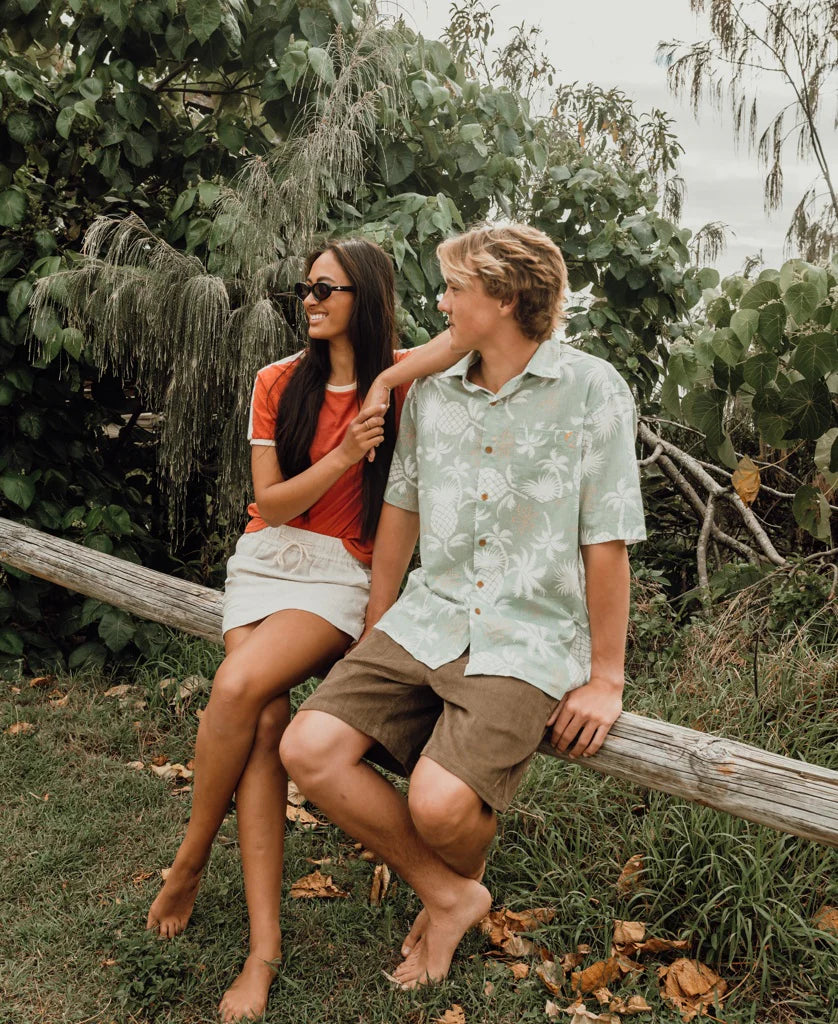 A woman wearing the Retro Ringer Tee | Wash Red Beige sits on a wooden log outdoors next to a man in a light green patterned short sleeve shirt and brown shorts, surrounded by lush greenery.