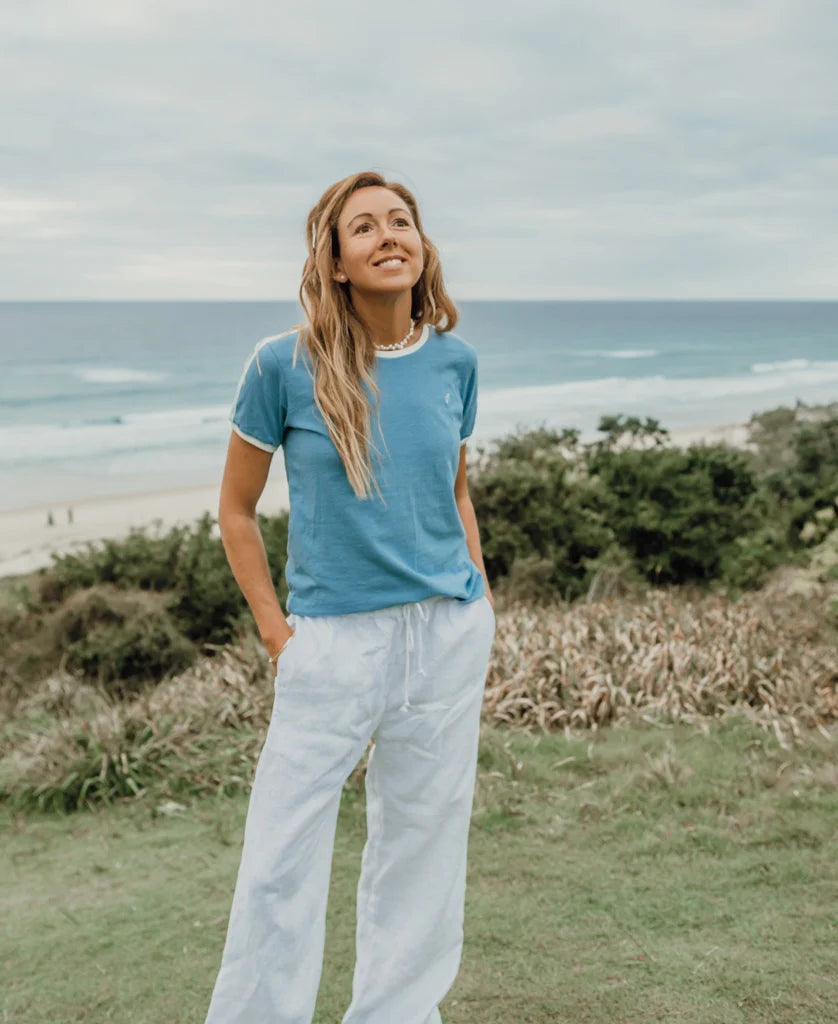 Woman standing near a beach wearing a Retro Ringer Tee | Azure with white pants, looking relaxed and enjoying the coastal view in a casual surfwear style.