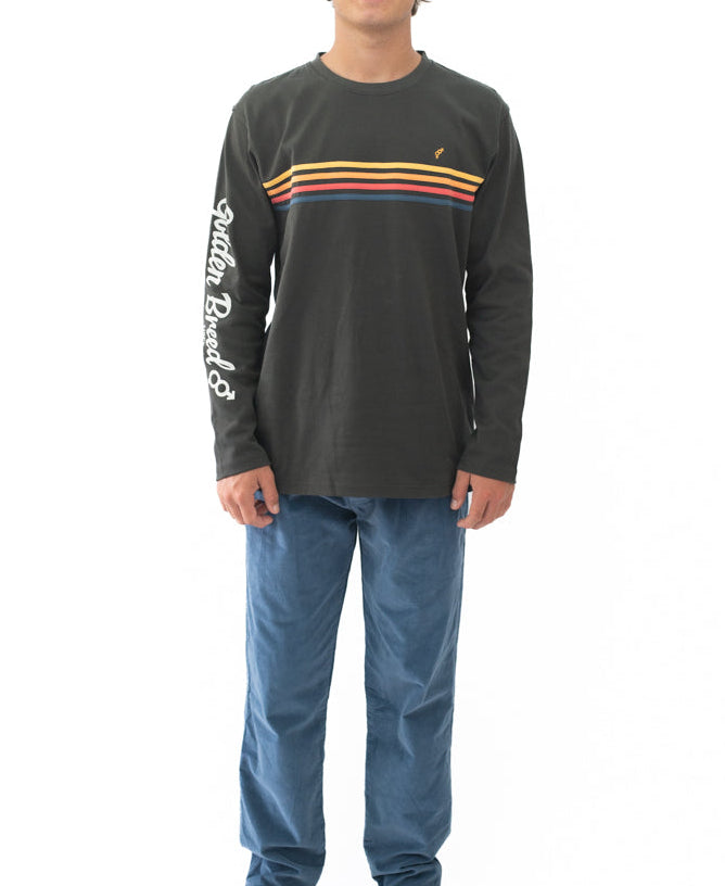Model wearing Heavy Island Long Sleeve Tee | Wash Black with yellow, red, and blue stripes across the chest and white script lettering on the right sleeve, paired with blue pants.