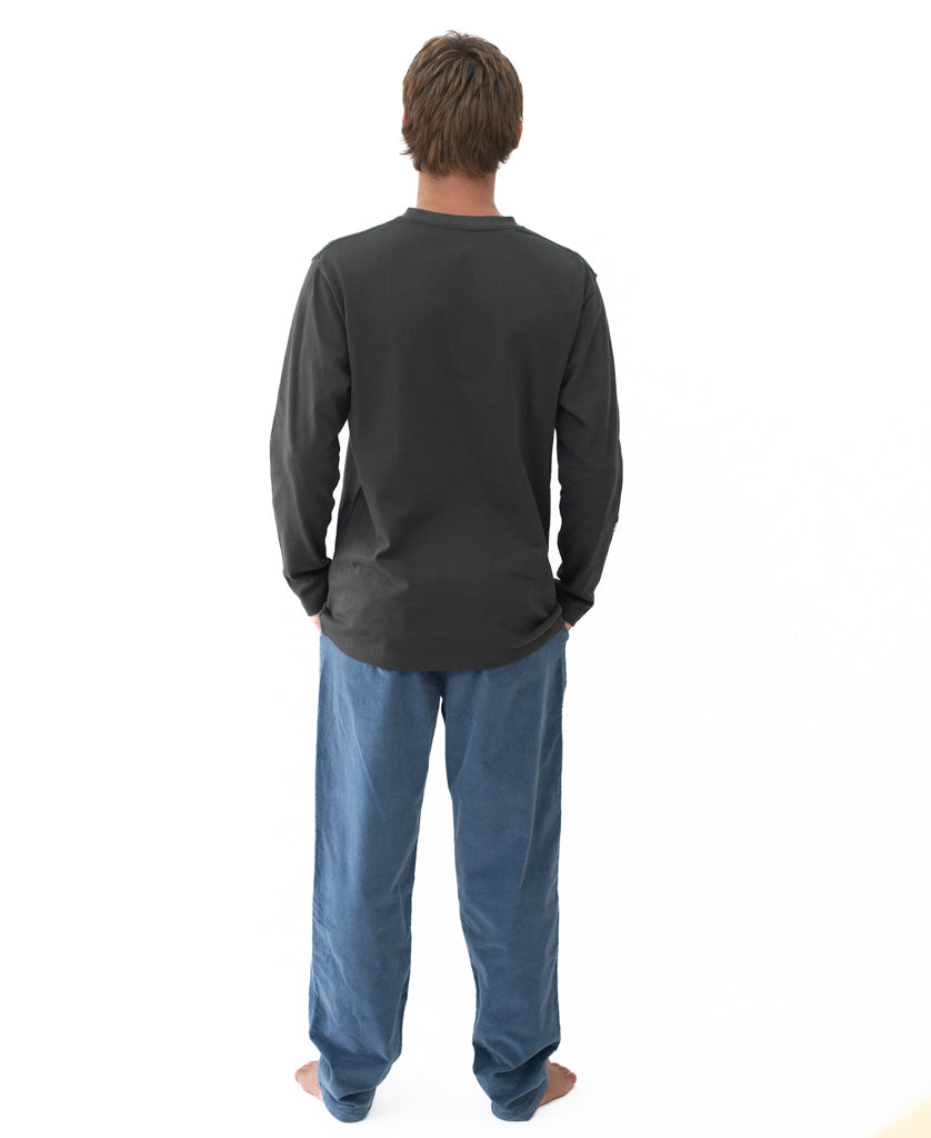 Man seen from behind wearing a Heavy Island Long Sleeve Tee | Wash Black paired with loose blue pants, standing barefoot against a plain white background.