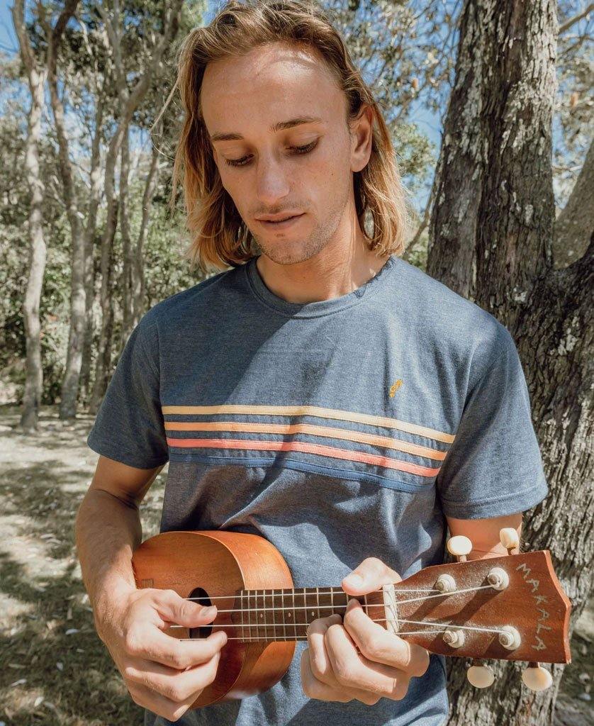Wearing the Golden Breed Long island Tee in Navy Marle, a person with long hair plays a wooden ukulele outdoors near trees, capturing a laid-back California surf style.