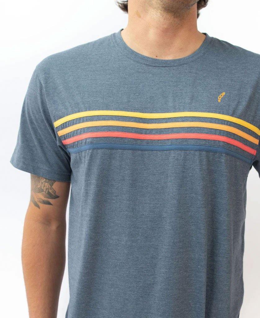 A person wears the Golden Breed Long Island Tee in Navy Marle, featuring yellow, orange, and red stripes across the chest for a California surf vibe, standing against a plain light background.