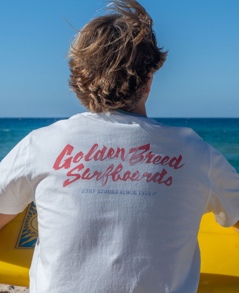 Back view of person in off-white surf tee featuring red Golden Breed Surfboards script logo, holding yellow board against ocean.